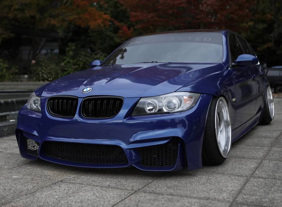 Lowered blue BMW 3 Series sedan fitted with M4 Look front bumper and large alloy wheels parked on pavement.
