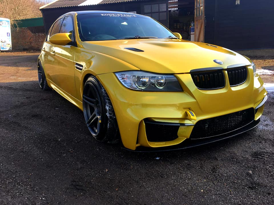Gold BMW 3 Series sedan with M4 Look front bumper, wide fenders, and black sport wheels parked outdoors.