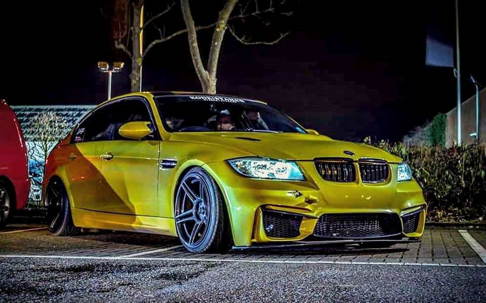 Gold BMW 3 Series E90 with M4 Look front bumper and custom wheels parked at night under artificial lights.