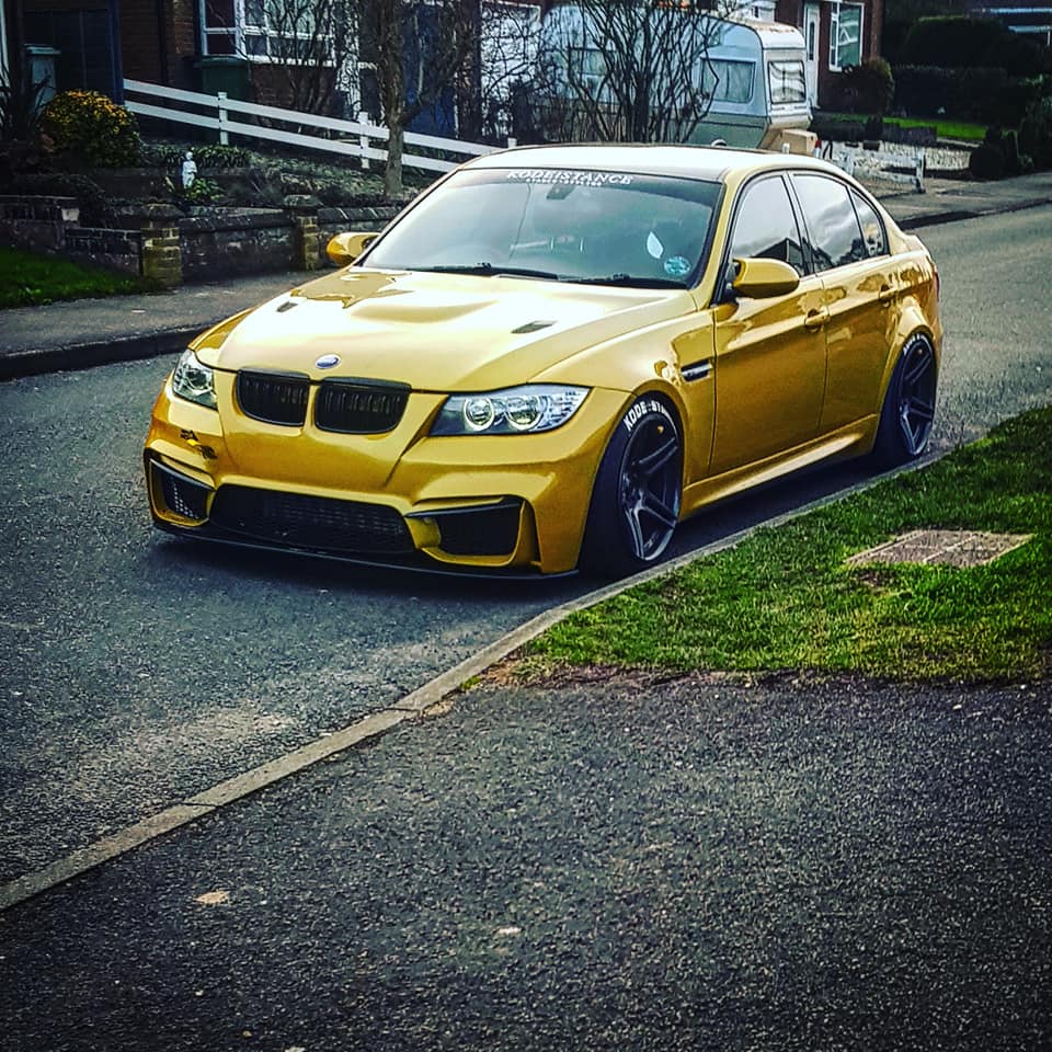 Gold BMW 3 Series sedan lowered with M4 Look front bumper and wide tires parked on residential street.
