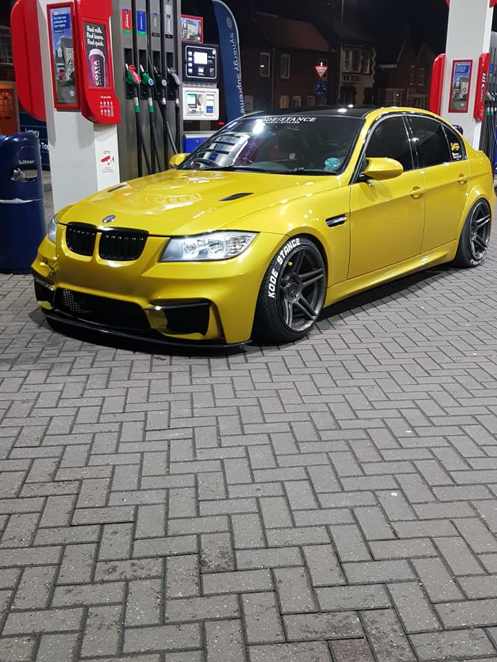 Gold BMW 3 Series sedan with M4 Look front bumper and sport wheels parked at a gas station.