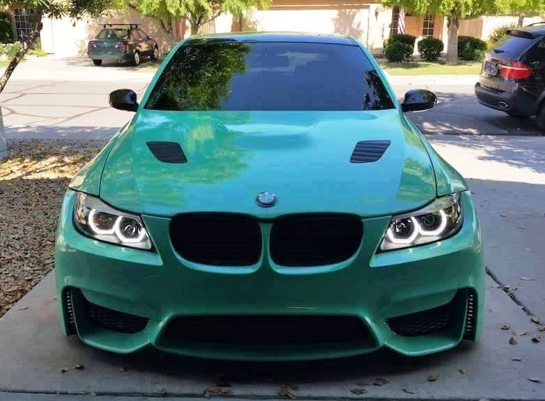 Green BMW 3 Series E90 with M4 Look front bumper, vented hood, and LED headlights parked in driveway.