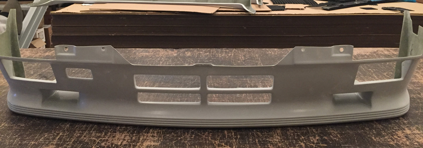 Front view of a Hartge H23 look front bumper add-on for BMW E30 PH1, displayed on a wooden surface.