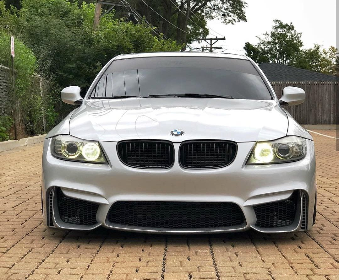 Front view of a silver BMW 3 Series E90 with M4 Look front bumper, wide air intakes, and headlights on.
