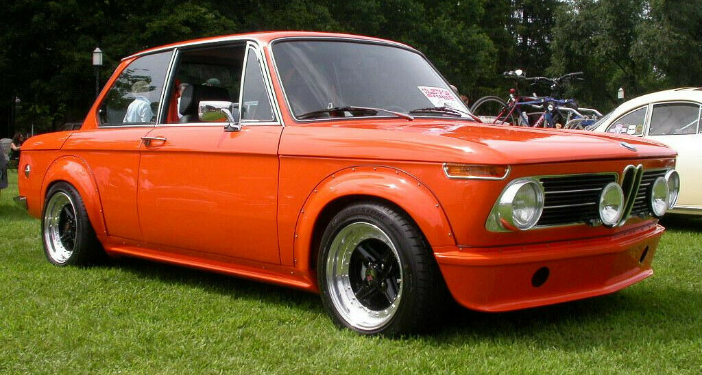 Orange BMW 2002 fitted with a front lip spoiler, aftermarket wheels, and rally-style headlights, parked on grass.