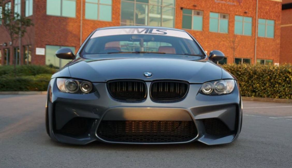 Grey BMW 3 Series E92 coupe with M2 look front bumper, aggressive design and large air intakes, parked outdoors.