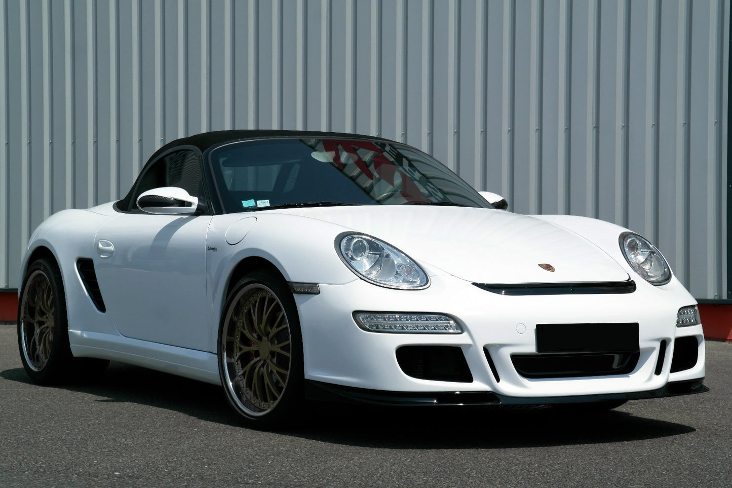 White Porsche Boxster 987 with GT3 Look front bumper, featuring large air intakes, LED daytime running lights, gold alloy wheels, and a black convertible roof.