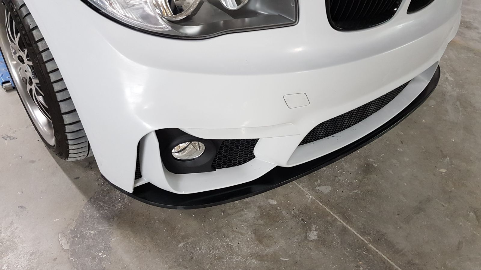Close-up of a BMW 1 Series E81/E82/E87/E88 front bumper with black M4 style lip installed.